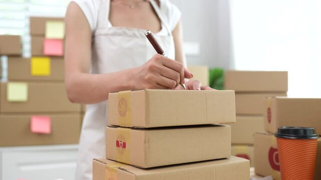 Young Asian businesswoman packing online orders at home office, using laptop for e-commerce sales and shipping in a small business.