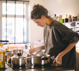 Woman standing by the stove in the kitchen, cooking and smelling the nice aromas from her meal in a pot. Housewife cooking lunch or breakfast.
