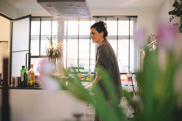 Cheerful woman cooking in a bright kitchen, surrounded by natural light and green plants. Candid lifestyle moment with soft focus foreground, homey interior, and relaxed morning routine.