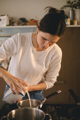 Young woman in casual clothes pouring milk into a saucepan pot while cooking at home. Natural kitchen setting, candid moment, warm lighting, everyday lifestyle, simple and real domestic atmosphere.