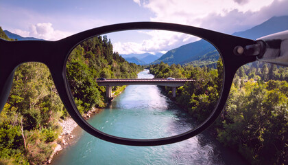 Eyeglasses frame a river scene with a bridge mountains and lush greenery.