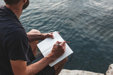 man sits on a rocky shore by water, writing in notebook. he is immersed in his thoughts, journaling...