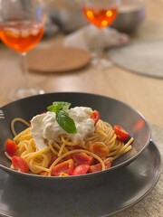 Spaghetti topped with stracciatella, cherry tomatoes, and fresh basil in black bowl near aperol spritz cocktails, highlighting italian cuisine