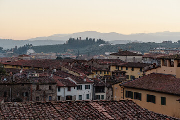 Sunrise view of rustic Tuscan village rooftops with a distant, scenic countryside featuring trees and a castle on the hills, creating a serene and charming landscape