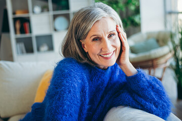 Charming elderly woman smiling indoors wearing a cozy blue sweater, relaxing in a beautifully...