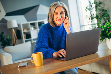 Happy mature woman working on laptop at home with a cup of coffee, enjoying casual and comfortable...