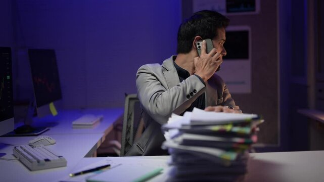 A man is sitting at a desk with a computer monitor and a keyboard. He is talking on a cell phone. The room is dimly lit, and there are stacks of papers on the desk. The man is focused on his work