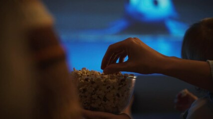 A hand reaches for popcorn from a container while a child sits nearby, both engaged in a cozy movie night at home. The dimly lit room creates a warm atmosphere as a film plays in the background
