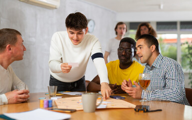 Emotional cheerful young guy playing interesting board game with male friends of different...