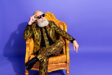 Stylish elder man in eccentric outfit seated on a golden armchair on a striking purple background