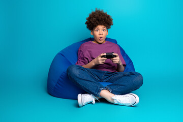 Young boy with curly hair playing a video game while sitting on a blue beanbag chair against a vibrant backdrop.