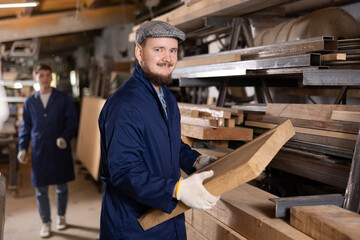 Young man worker takes wooden plank from stack in wood workshop