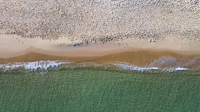 The sand looks brown and has a few visible footprints. The water is blue green, with waves crashing on the shore.