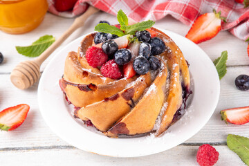 Sweet summer bundt cake with fresh fruit and berry. Traditional round sponge cake, seasonal dessert with strawberry, raspberry, blueberry, honey and sugar sprinkles, white wooden table copy space