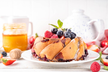 Sweet summer bundt cake with fresh fruit and berry. Traditional round sponge cake, seasonal dessert with strawberry, raspberry, blueberry, honey and sugar sprinkles, white wooden table copy space