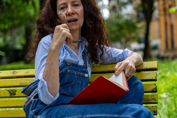 middle aged woman sitting on a yellow bench looking into the camera reading a book with a red cover