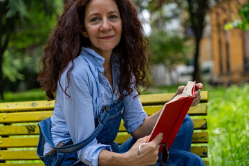 middle-aged woman with red hair sitting on a yellow bench reading a book