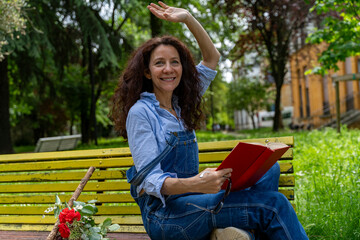 middle aged woman sitting on a yellow bench waving while reading a book
