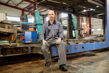Portrait of a senior Caucasian mechanic sitting in an industrial workshop with machinery and tools...
