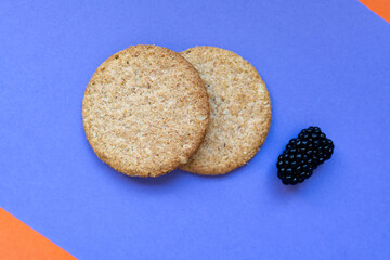 Two round oatmeal cookies and fresh blackberries on a vibrant geometric background in orange and purple tones. Minimalist style with soft focus and summer vibes.