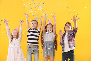 Happy children in conical paper hats under falling confetti on yellow background. Birthday surprise...