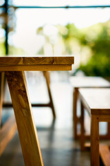 Closeup of a rustic wooden table edge and sturdy leg, showcasing natural grain and craftsmanship. Bright, blurred outdoor background with greenery evokes a warm, inviting al fresco dining vibe.