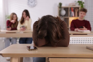 Cyber bullying. Upset teenage girl with smartphone and schoolmates pointing at her in classroom,...
