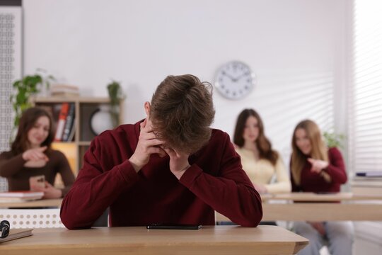 Cyber bullying. Upset teenage boy with smartphone and schoolmates pointing at him in classroom, selective focus