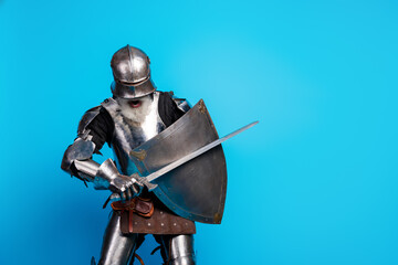Elderly man in medieval knight armor posing on blue background with sword and shield, showcasing...