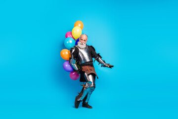 Senior man in medieval armor holding colorful balloons against blue background portraying heroism...