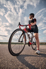 Low angle view photo of woman standing next to racing bike, one foot forward, glancing into distance, sky filled with soft clouds in background. Concept of sport training, vision, ambition, rest.