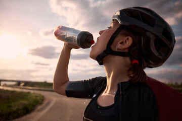Close-up photo of female cyclist pouring water from bottle into mouth, sunlight behind and road...
