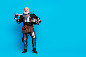 Elderly man dressed in ornate knight armor holding a sword against a blue studio background
