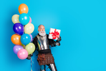Mature man dressed as a medieval knight holding colorful balloons and a gift box against blue...