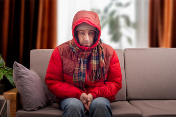Middle age male model in red parka and checkered scarf getting cold sitting on the sofa in cold living room