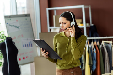 Young female fashion designer working in modern atelier with clipboard while multitasking and...