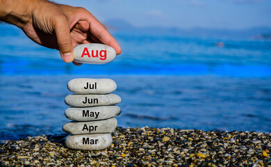 A hand places a stone labeled "Aug" on top of a stacked tower of stones representing the months March through August at a beach.