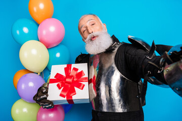 Festive senior man in knight armor holding gift box while posing near colorful balloons against...