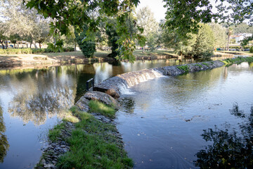 A serene river scene featuring a small stone dam with water gently cascading over the edge, surrounded by greenery and reflected sunlight, creating a peaceful and natural atmosphere. Serta, Portugal.