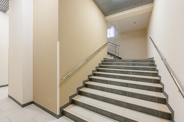 Fototapeta premium indoor staircase with tile steps and metal handrails. The walls are beige, and the ceiling has a grid pattern. There's a small window at the top of the stairs.