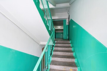 Stairwell with teal and white walls. Concrete stairs with teal railing lead upwards. A doorway is visible at the top of the stairs