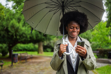 Smiling businesswoman using a smartphone while holding an umbrella in a rainy day in a park,...