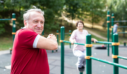 Obraz premium Staying after retirement. Happy joyful mature retired sportsman wearing sportswear doing side stretching exercises with arm over his head, exercising outside in city park