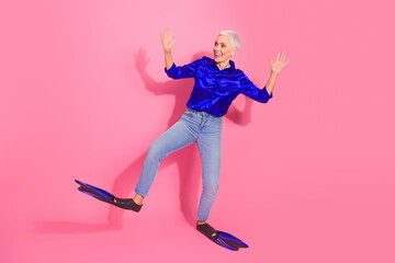 Cheerful mature woman in stylish blue shirt and jeans enjoying playful moment with pink background and flippers