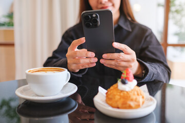 Closeup of a woman holding and using mobile phone with coffee cup and cake on the table in cafe