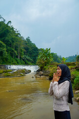Naklejka premium view of asian muslim woman expressing shock with hands in front of face facing river bank with waterfall background with clear sky for advertisement space.