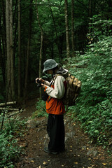 Young man hiking through dense green forest with camera in hand, wearing backpack and rain jacket, exploring nature and documenting.