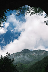 Dramatic view of cloudy sky and rocky mountain peaks framed by tree silhouettes, captured from within a cave on Mount Olympus.
