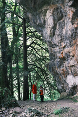 Two hikers in bright jackets stand beneath a towering rock formation in the lush, green forest of Mount Olympus, Greece.
