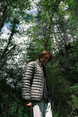A young man wearing a gray puffer jacket stands in a sunlit forest on Mount Olympus, surrounded by...